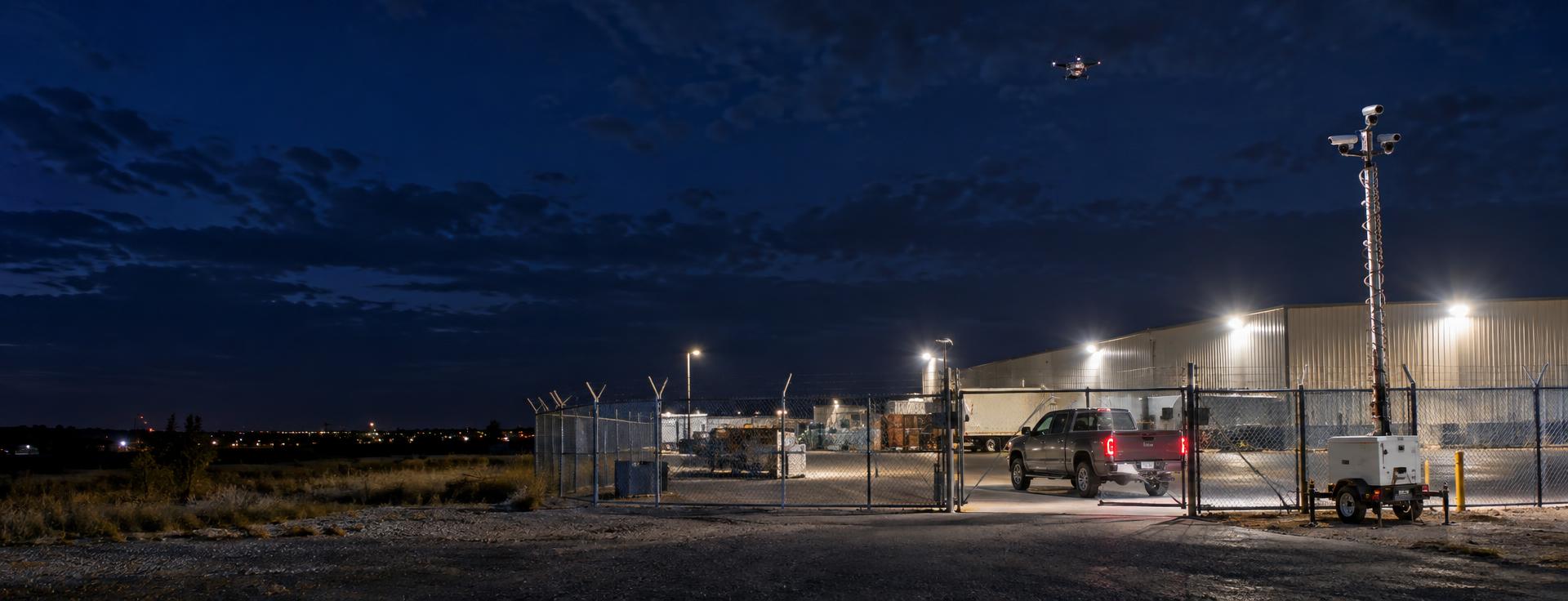 Security drone and mobile surveillance trailer documenting a vehicle at a fenced property gate at night